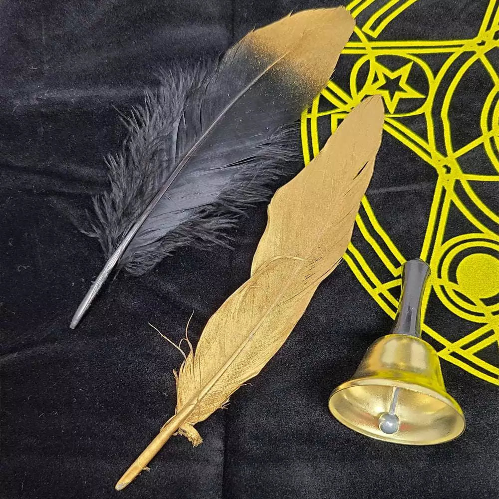 Black feather, golden quill, and small brass bell arranged on a dark surface with a yellow Rider-Waite mystical symbol in the background.