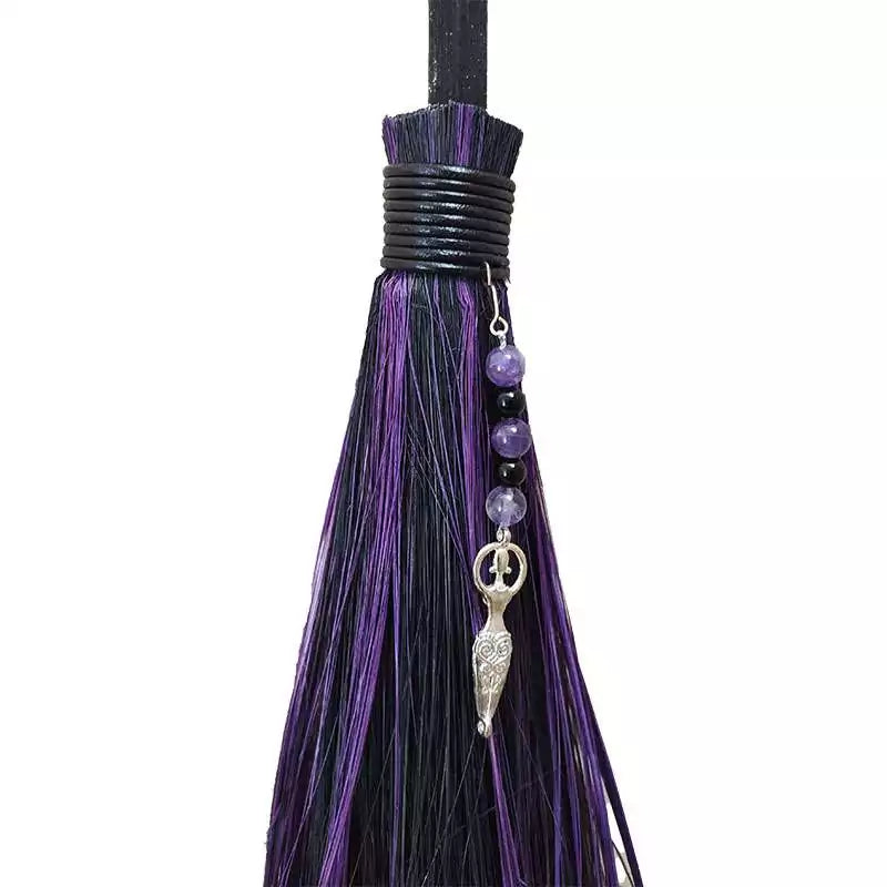Close-up of the decorative top of a besom broom showing purple and black tassels, small beads, and a silver charm pendant.
