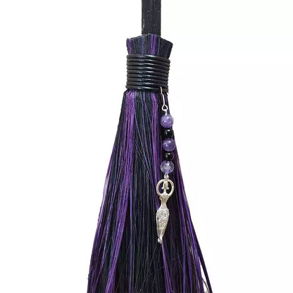 Close-up of the decorative top of a besom broom showing purple and black tassels, small beads, and a silver charm pendant.