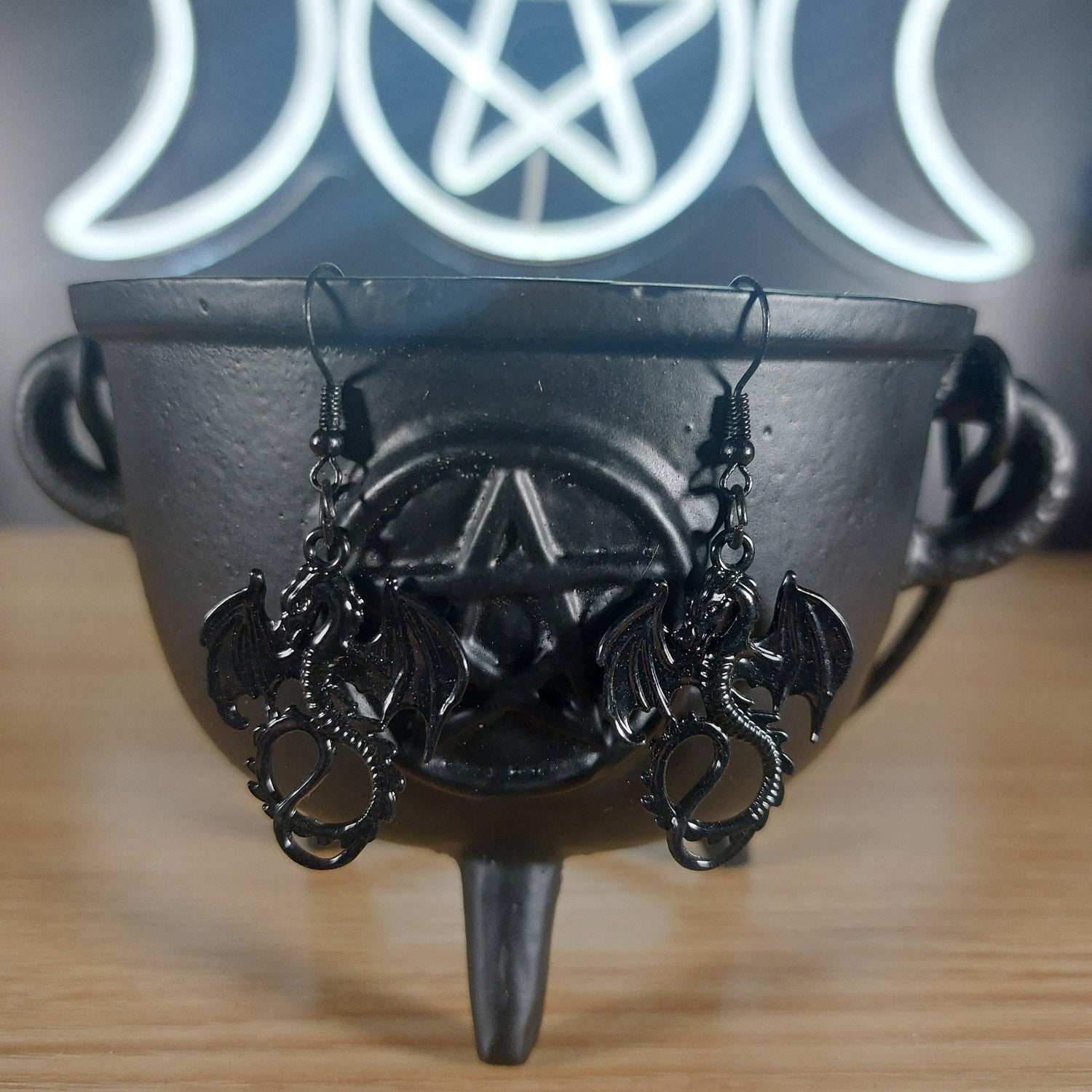 Black metal dragon dangle earrings with intricate wings, hanging from a black cauldron.