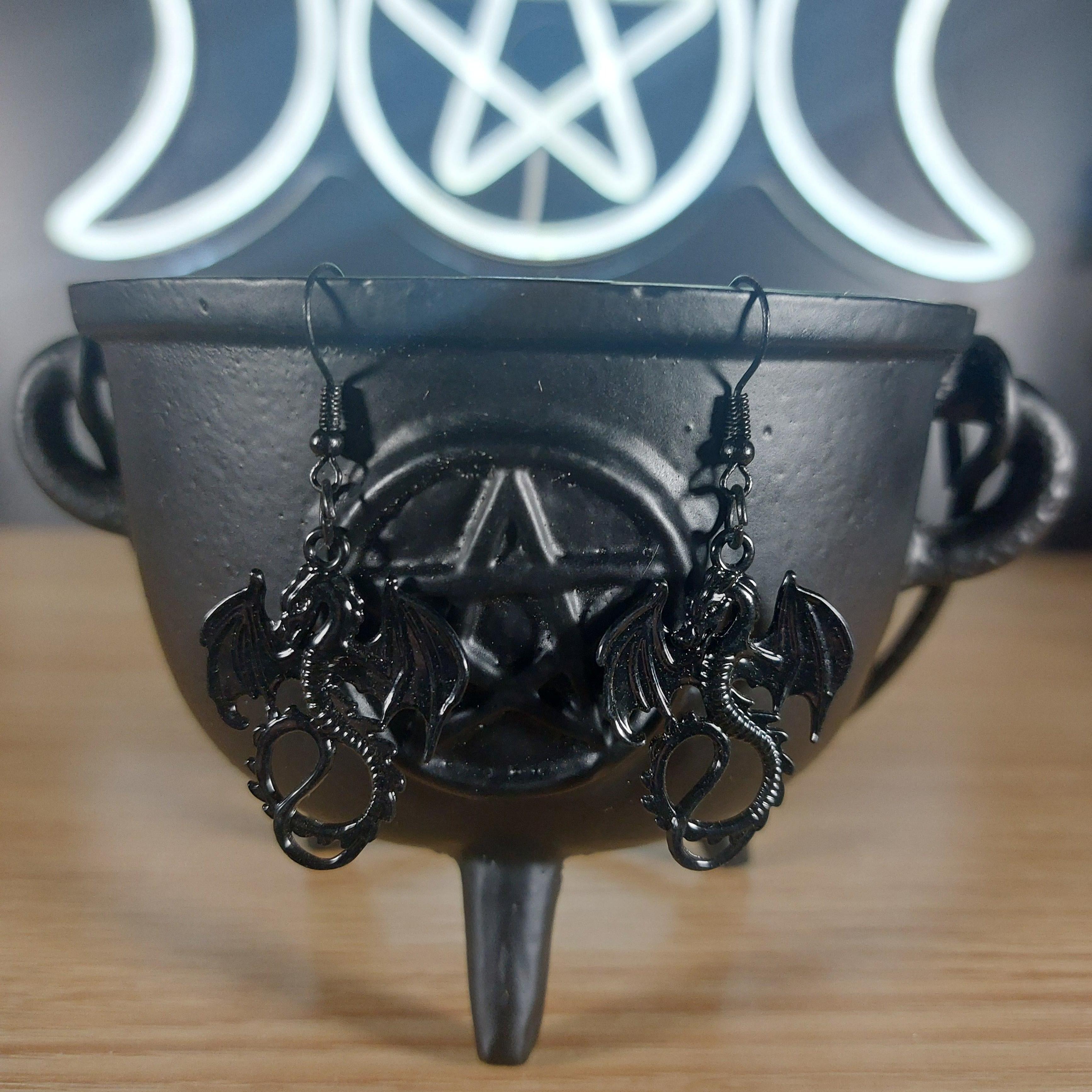 Black metal dragon dangle earrings with intricate wings, hanging from a black cauldron.