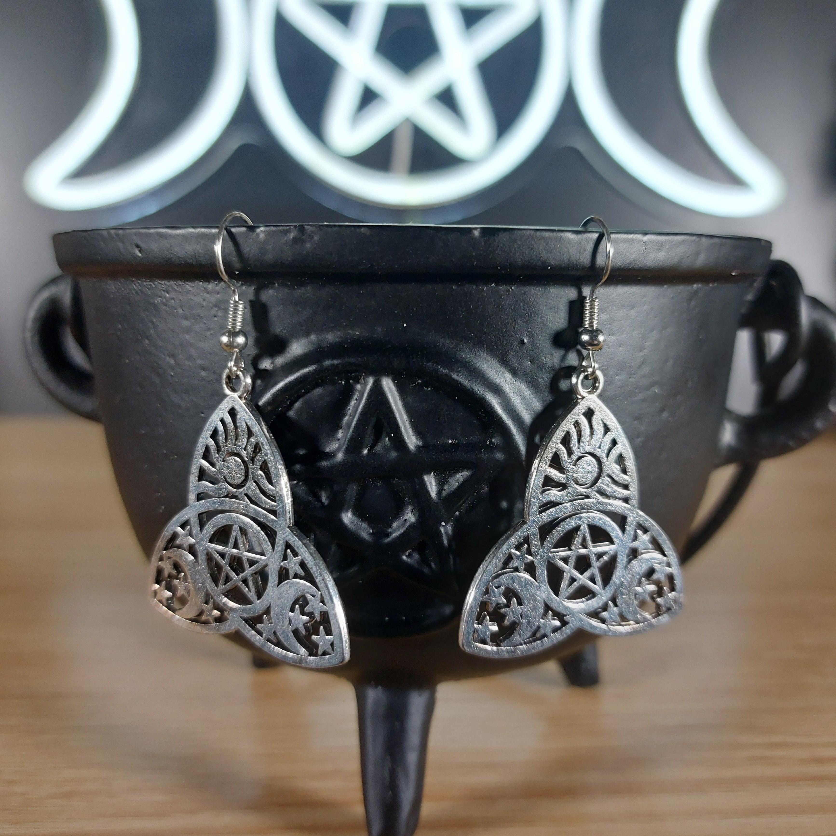 Silver tone triquetra dangle earrings with a pentagram design, displayed on a black cauldron.