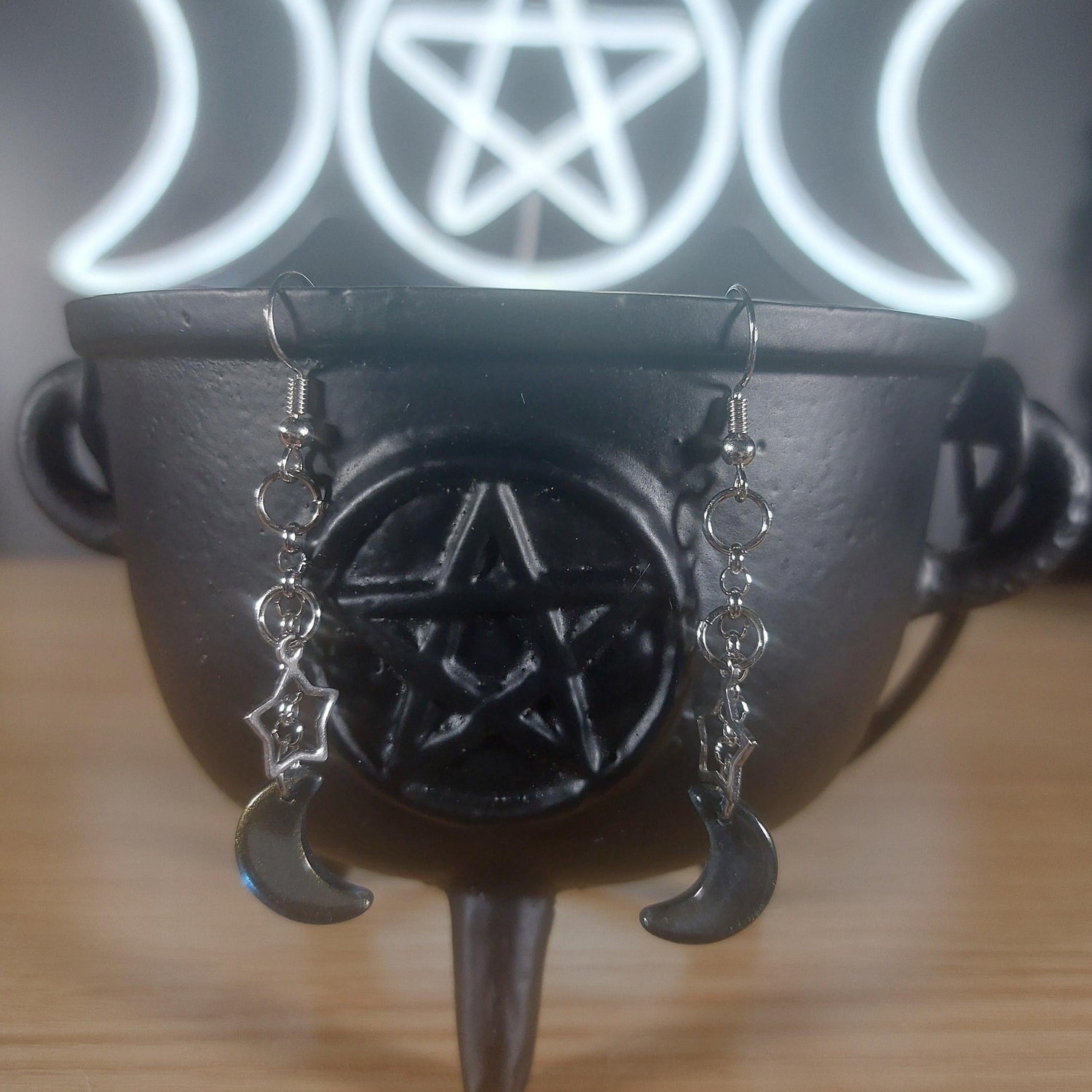 Silver moon and star dangle earrings hanging from a black cauldron, with a glowing triple moon and pentagram in the background.