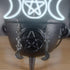Silver moon and star dangle earrings hanging from a black cauldron, with a glowing triple moon and pentagram in the background.