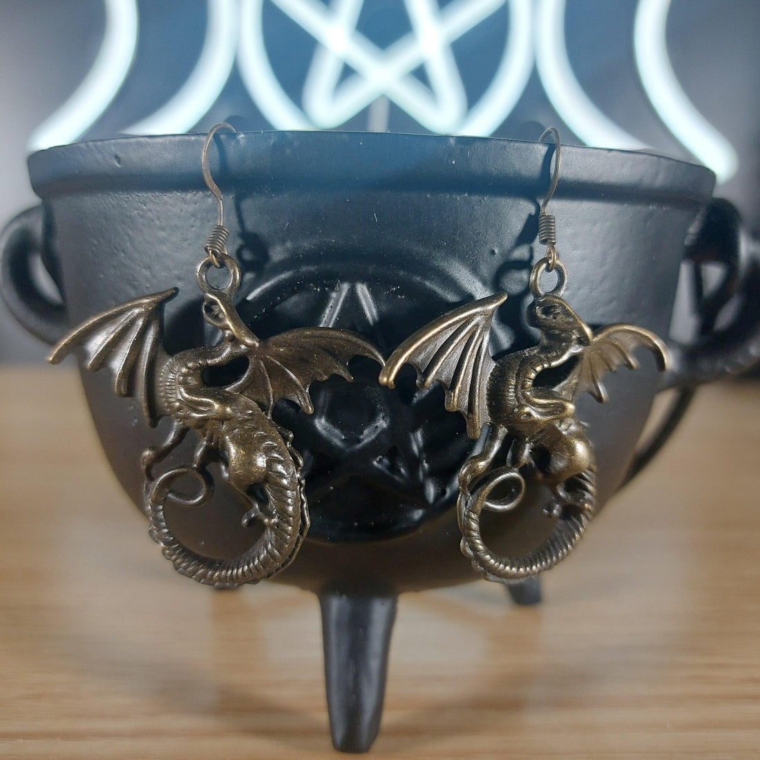 Pair of large bronze dragon dangle earrings with detailed wings and coiled bodies, hanging from a black cauldron.