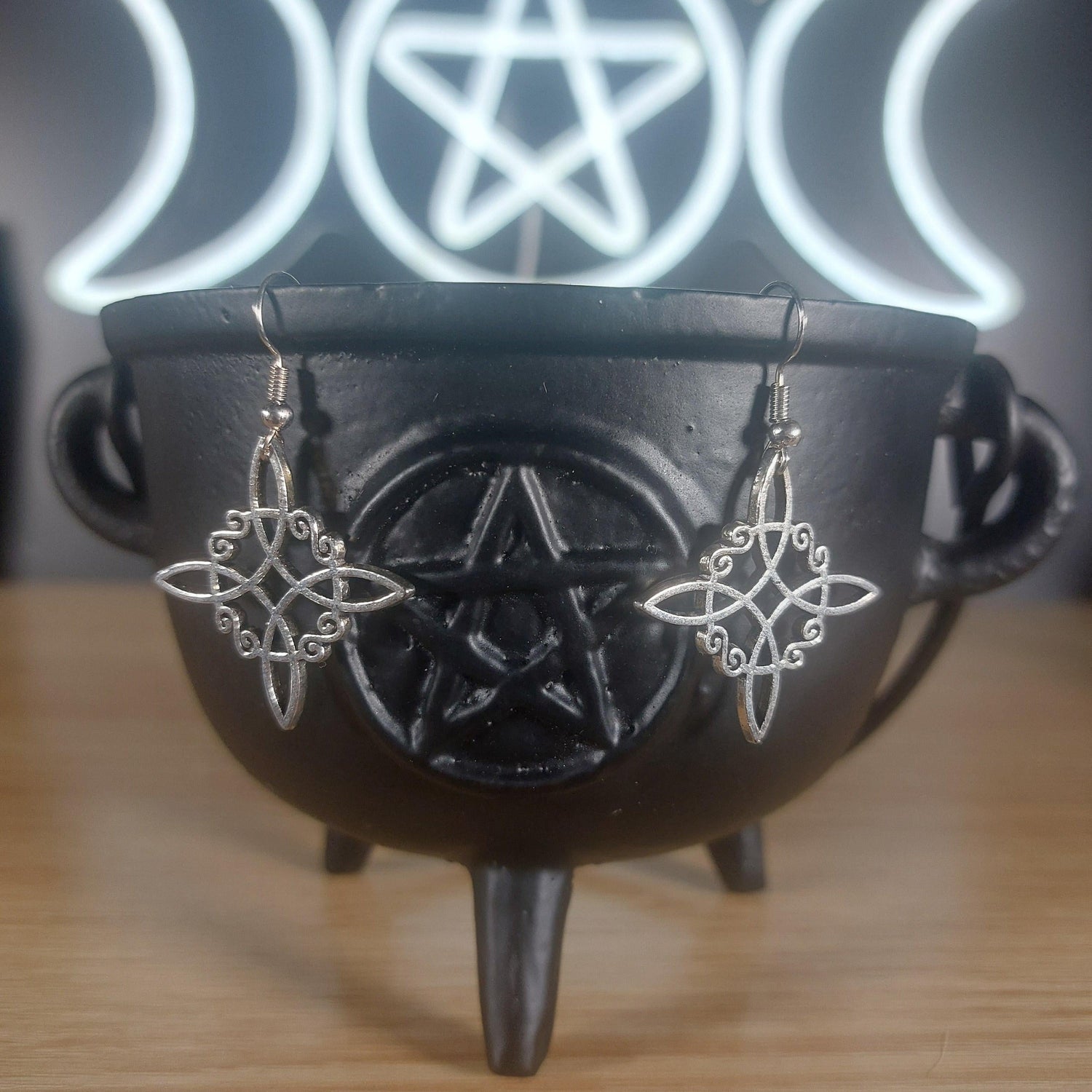Silver tone Witches Knot dangle earrings in a black cauldron, with a glowing Triple Moon and Pentagram symbol in the background.