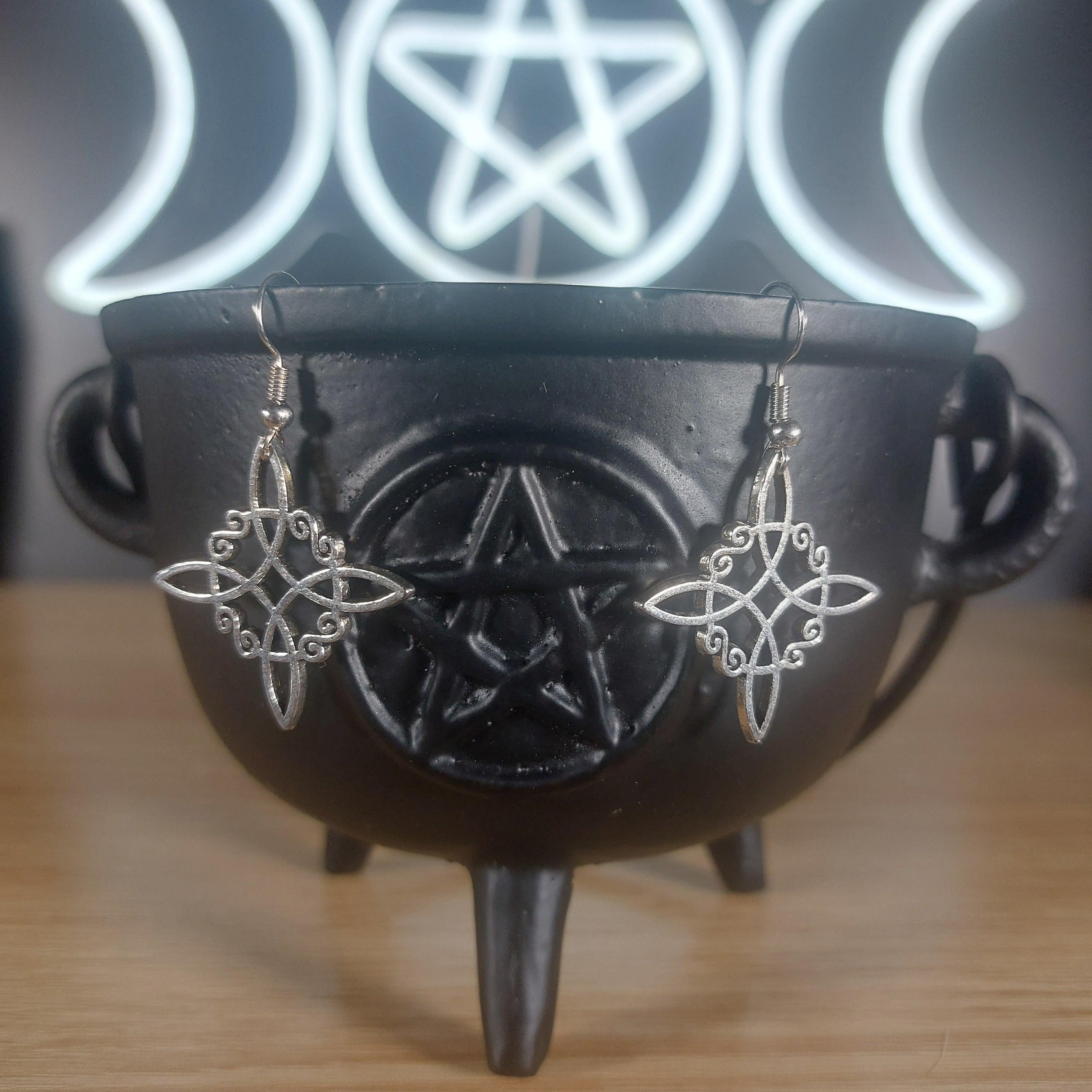 Silver tone Witches Knot dangle earrings in a black cauldron, with a glowing Triple Moon and Pentagram symbol in the background.