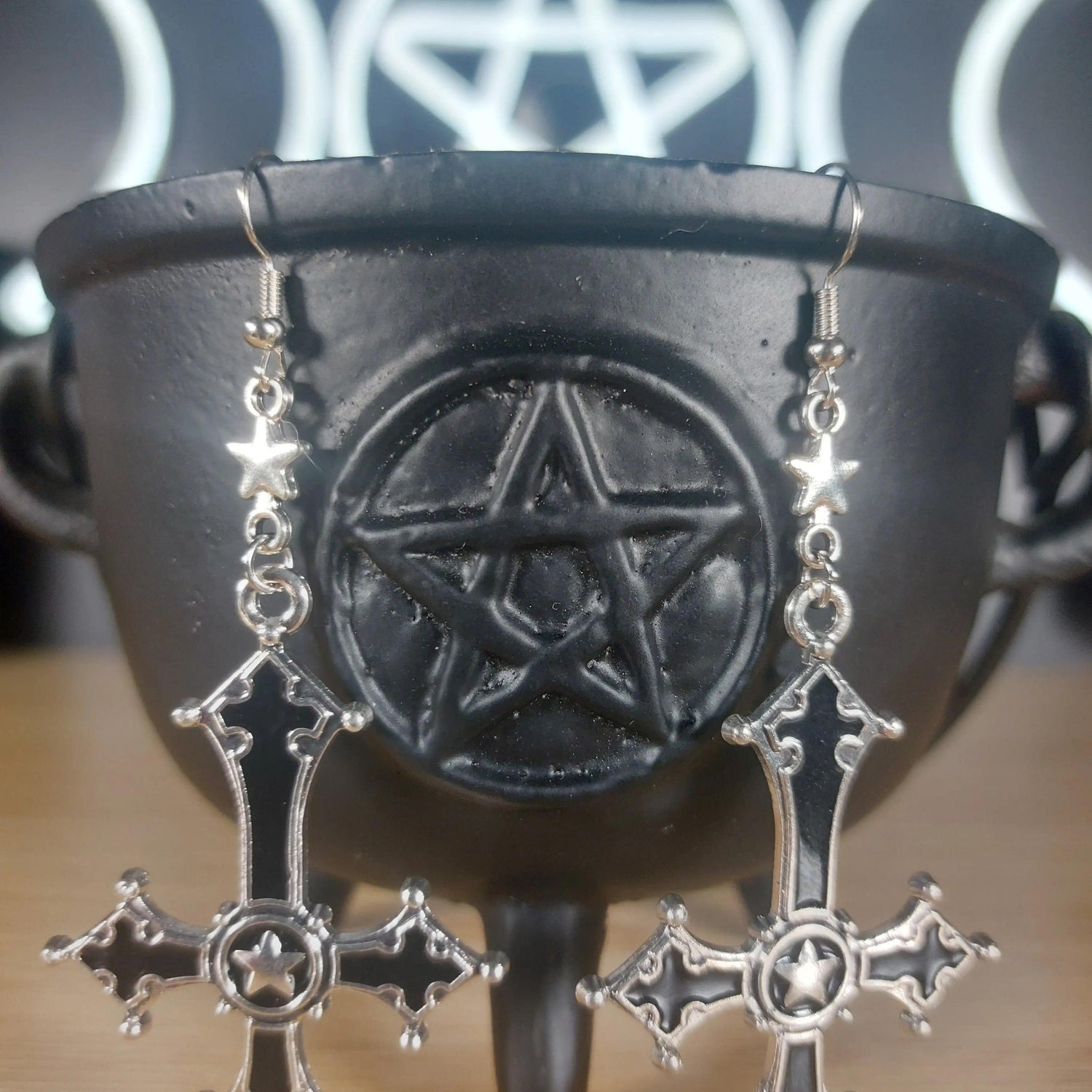 Silver and black upside-down cross earrings with star accents, hanging from a black cauldron.