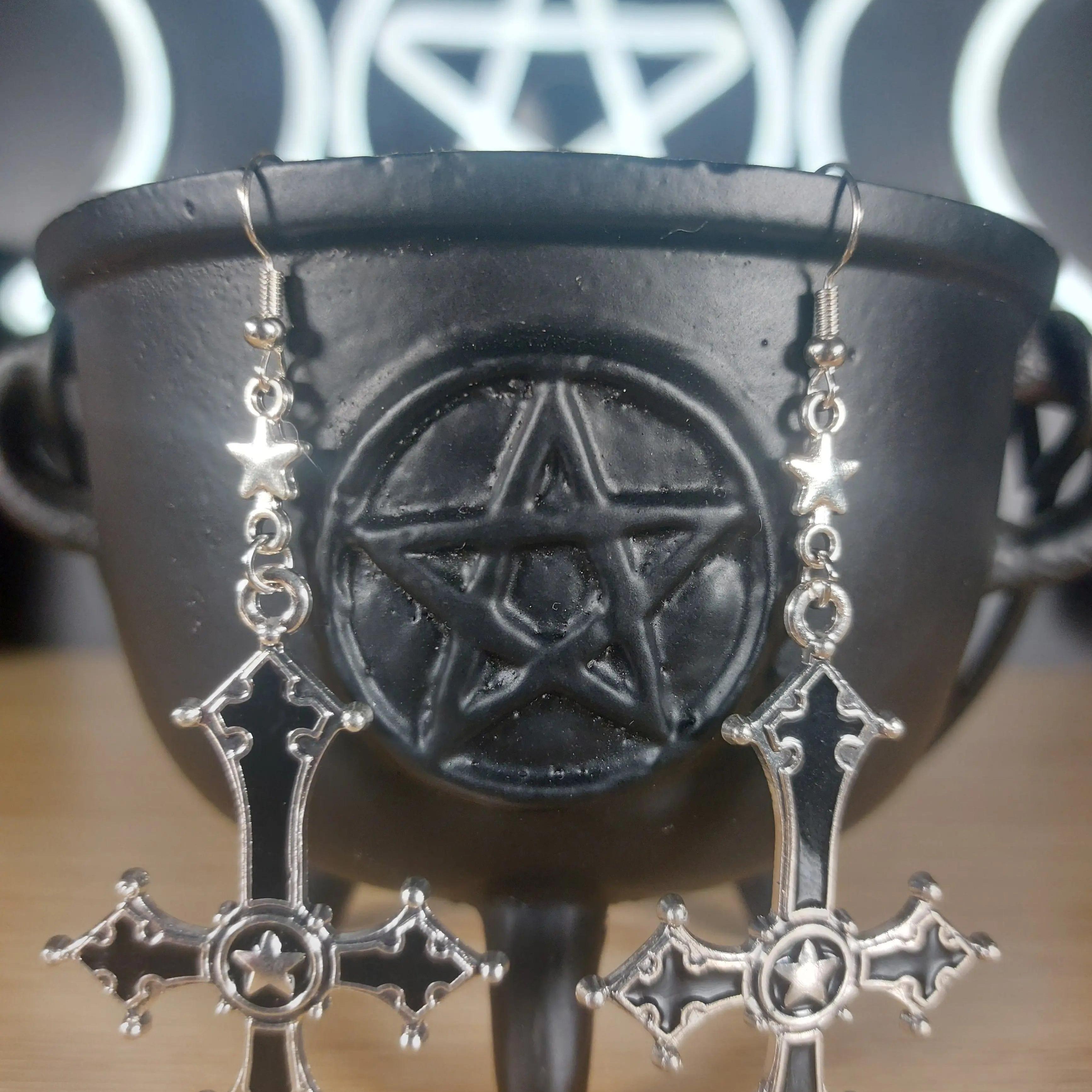 Silver and black upside-down cross earrings with star accents, hanging from a black cauldron.