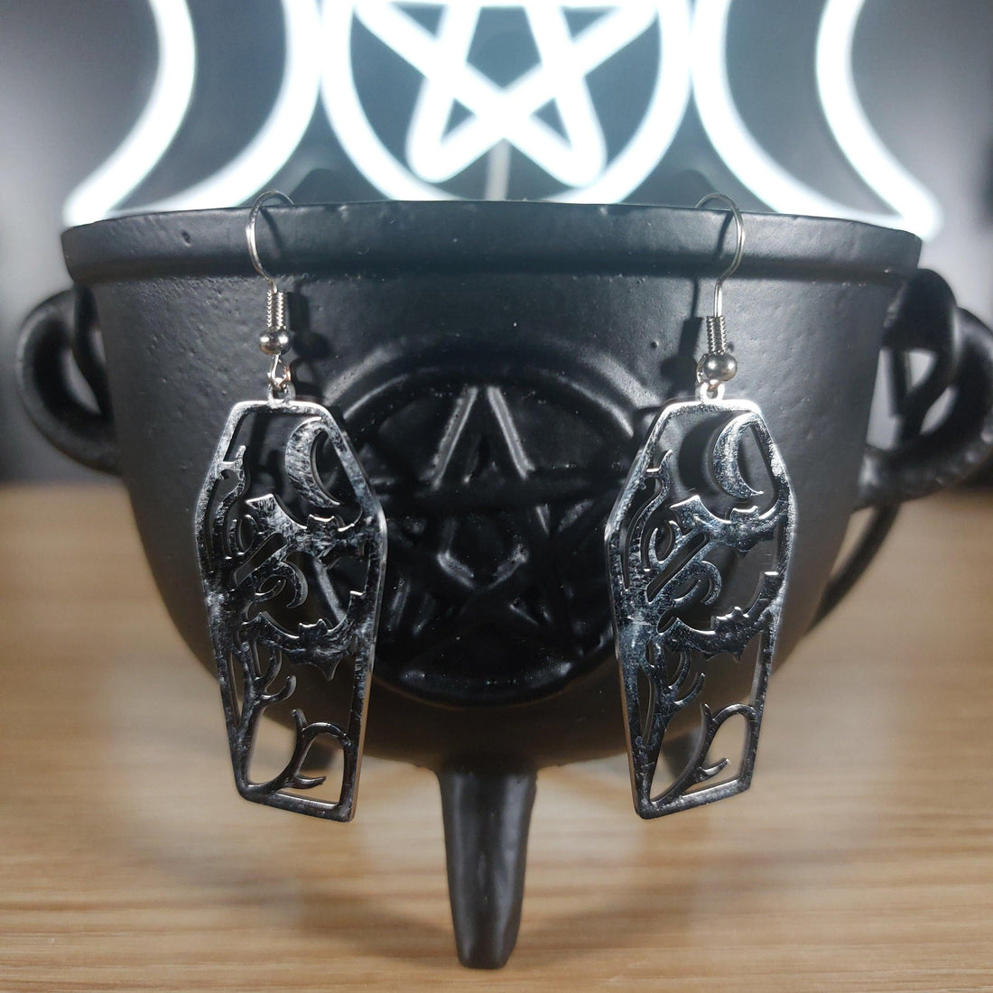 Silver tone coffin dangle earrings with cutout bats and moon design, hanging from a black cauldron.