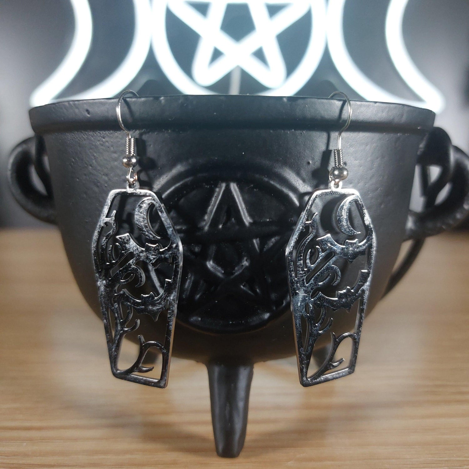 Silver tone coffin dangle earrings with cutout bats and moon design, hanging from a black cauldron.