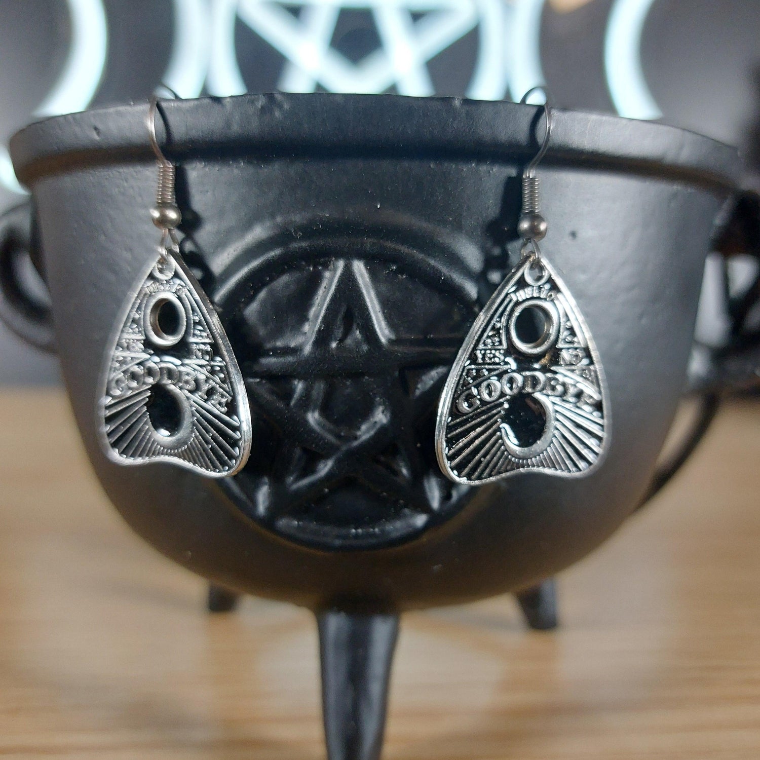 Silver tone Ouija planchette "Goodbye Yes No" dangle earrings on a black cauldron.