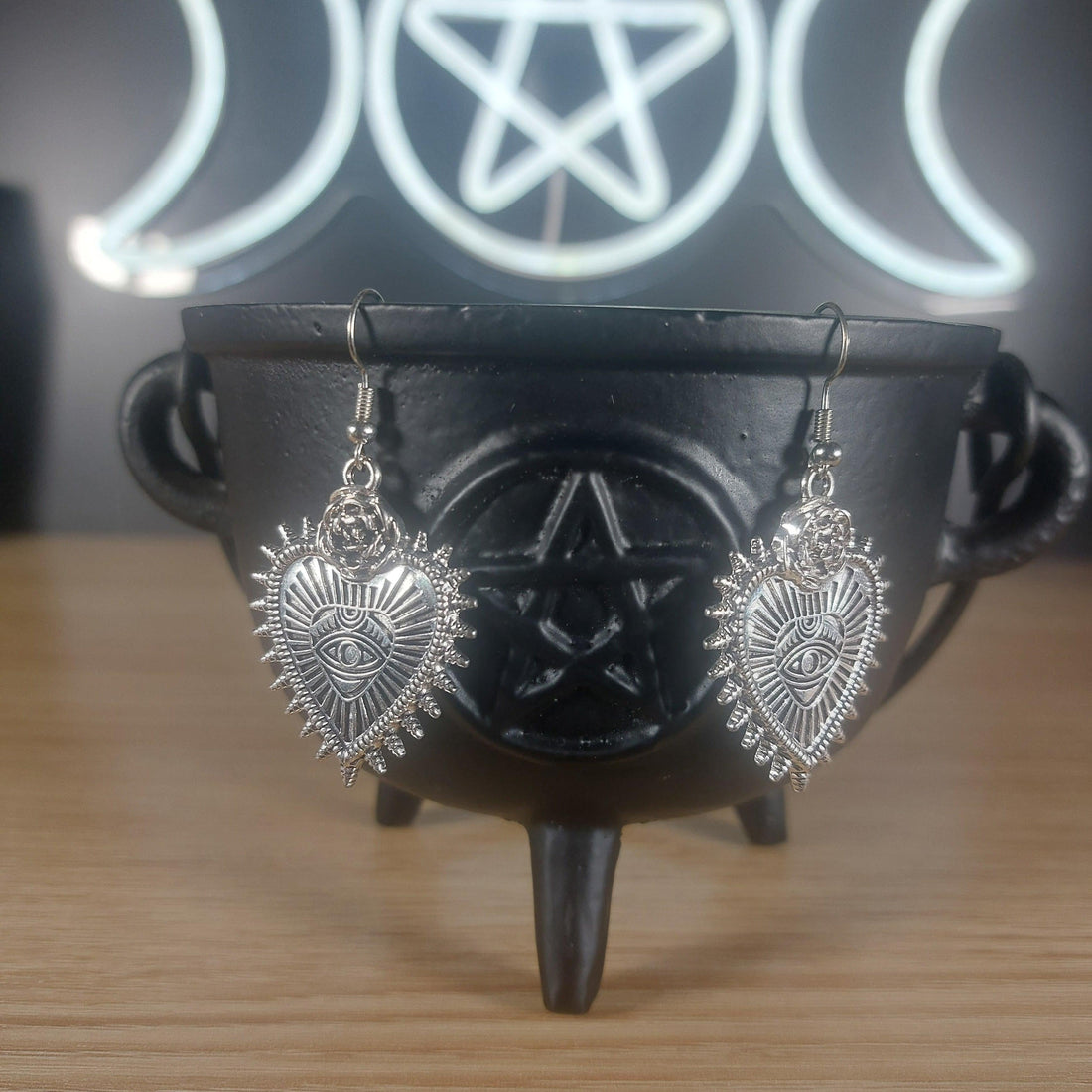 Silver evil eye heart dangle earrings hanging from a black cauldron, with a triple moon and pentagram neon sign in the background.