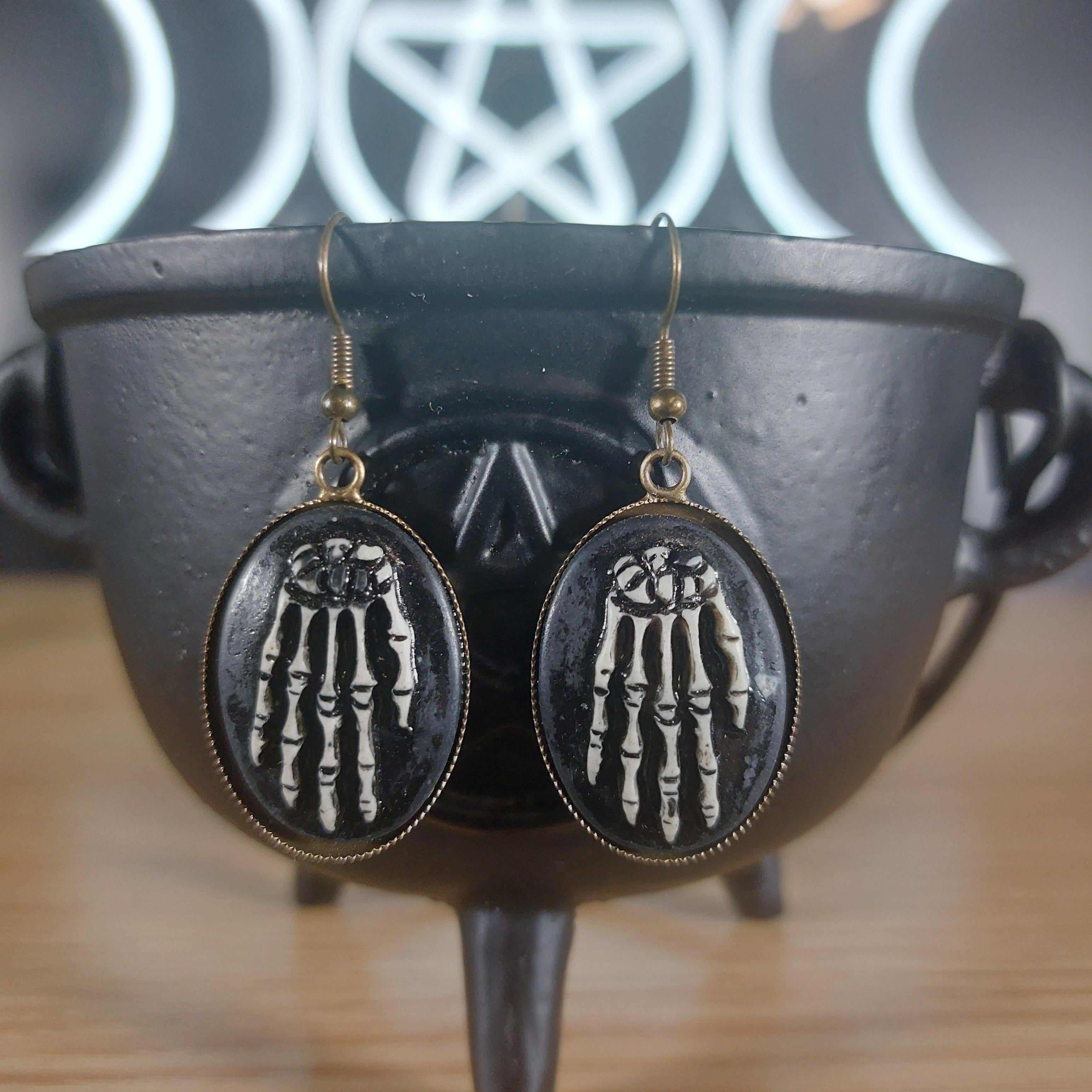 Antique bronze skeleton hands dangle earrings with black oval frames, resting on a black cauldron.