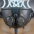 Antique bronze skeleton hands dangle earrings with black oval frames, resting on a black cauldron.