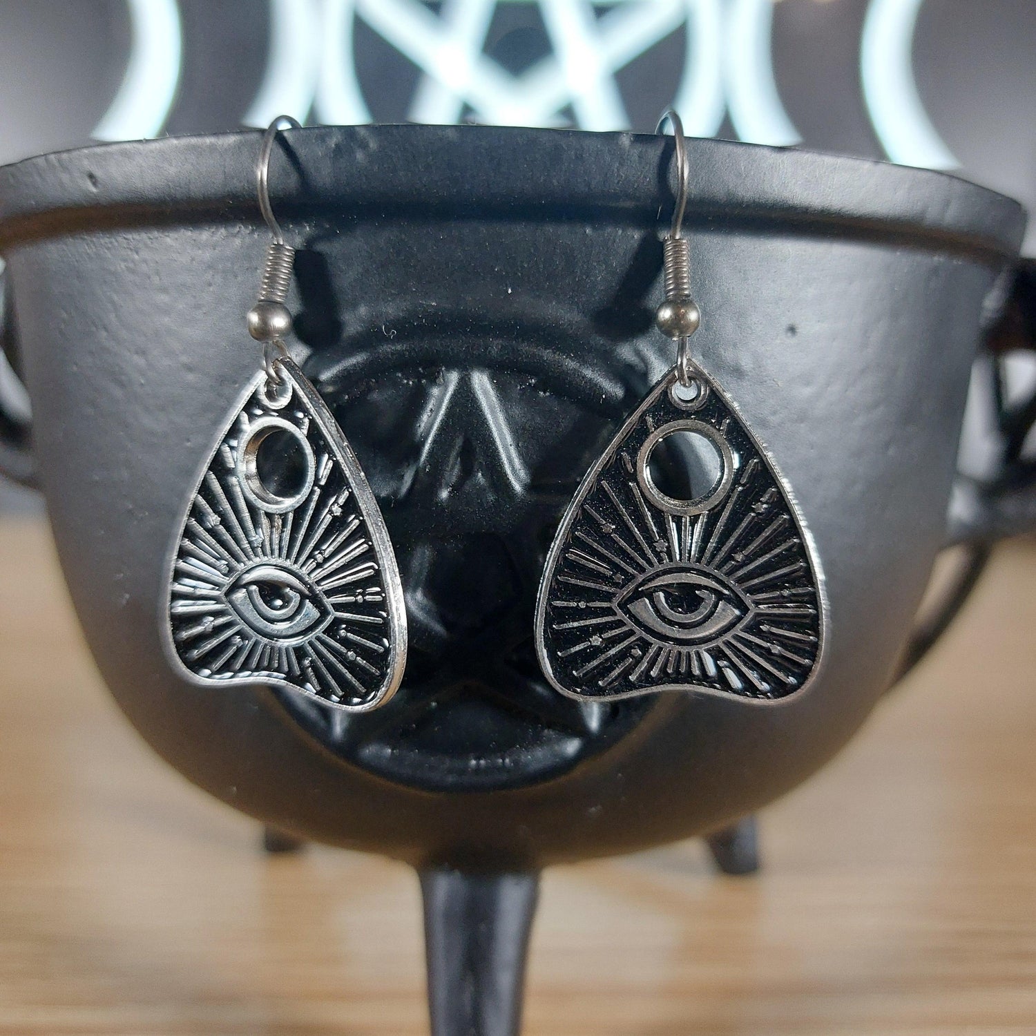 Silver tone evil eye quija planchette dangle earrings hanging from a black cauldron.