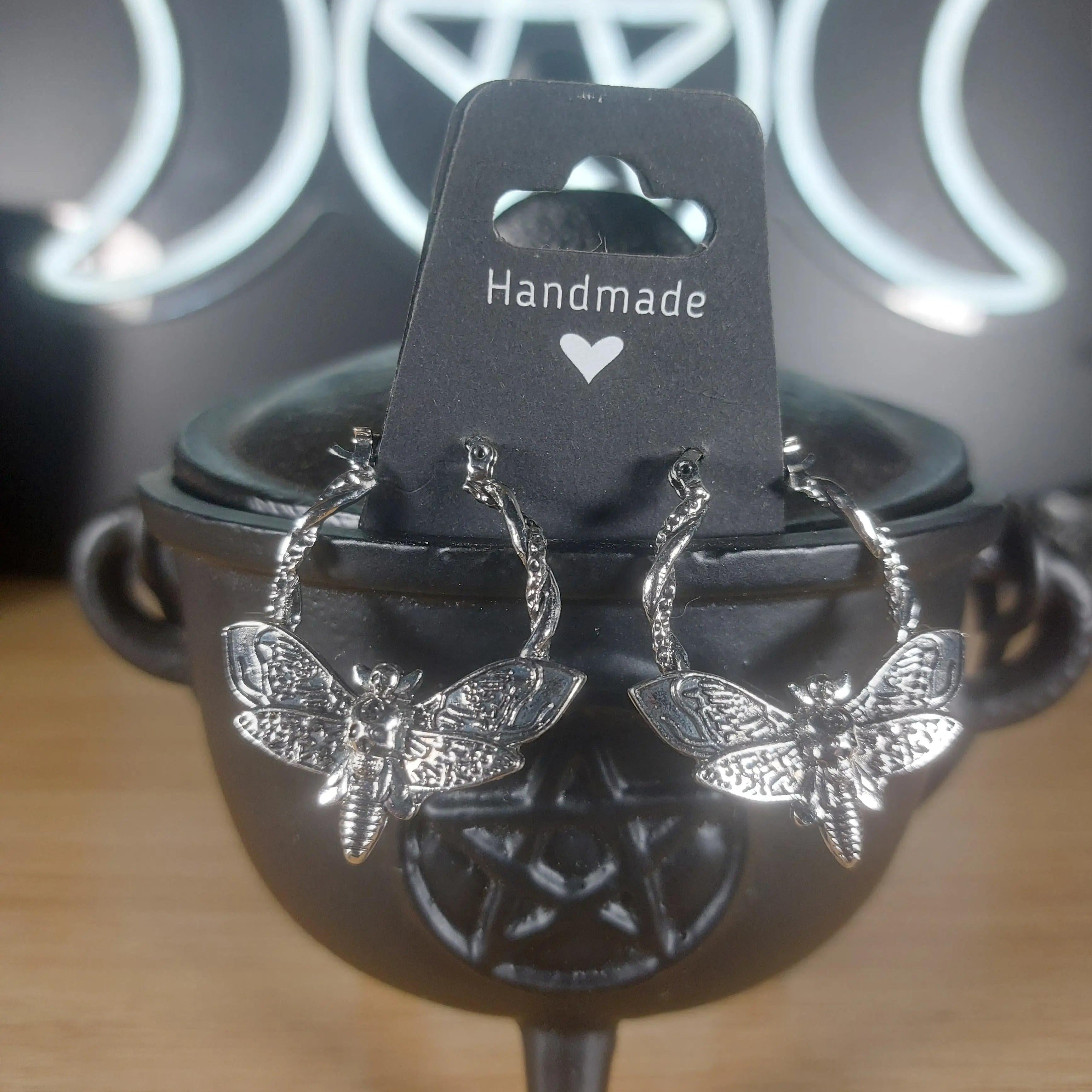 Silver tone death moth hoop earrings on a black "Handmade" card, displayed on a black cauldron.