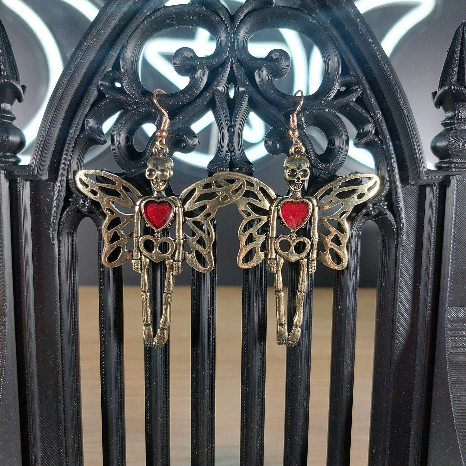 Gold tone skeleton with wings and red heart dangle earrings on a black gothic arch.