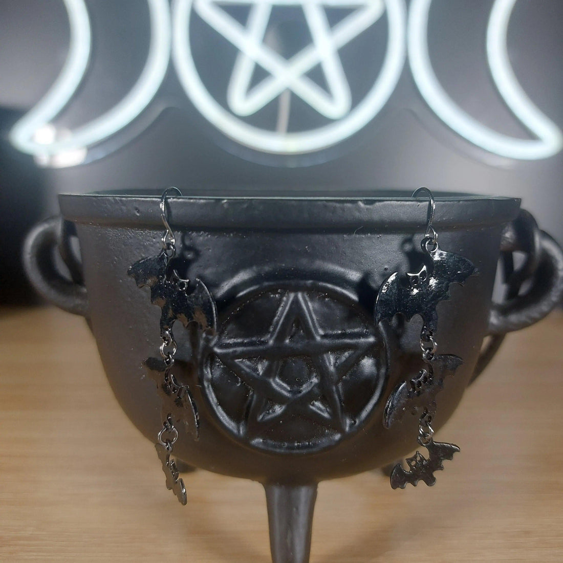 Black metal 3-tier bat dangle earrings hanging from a black cauldron with a pentagram.