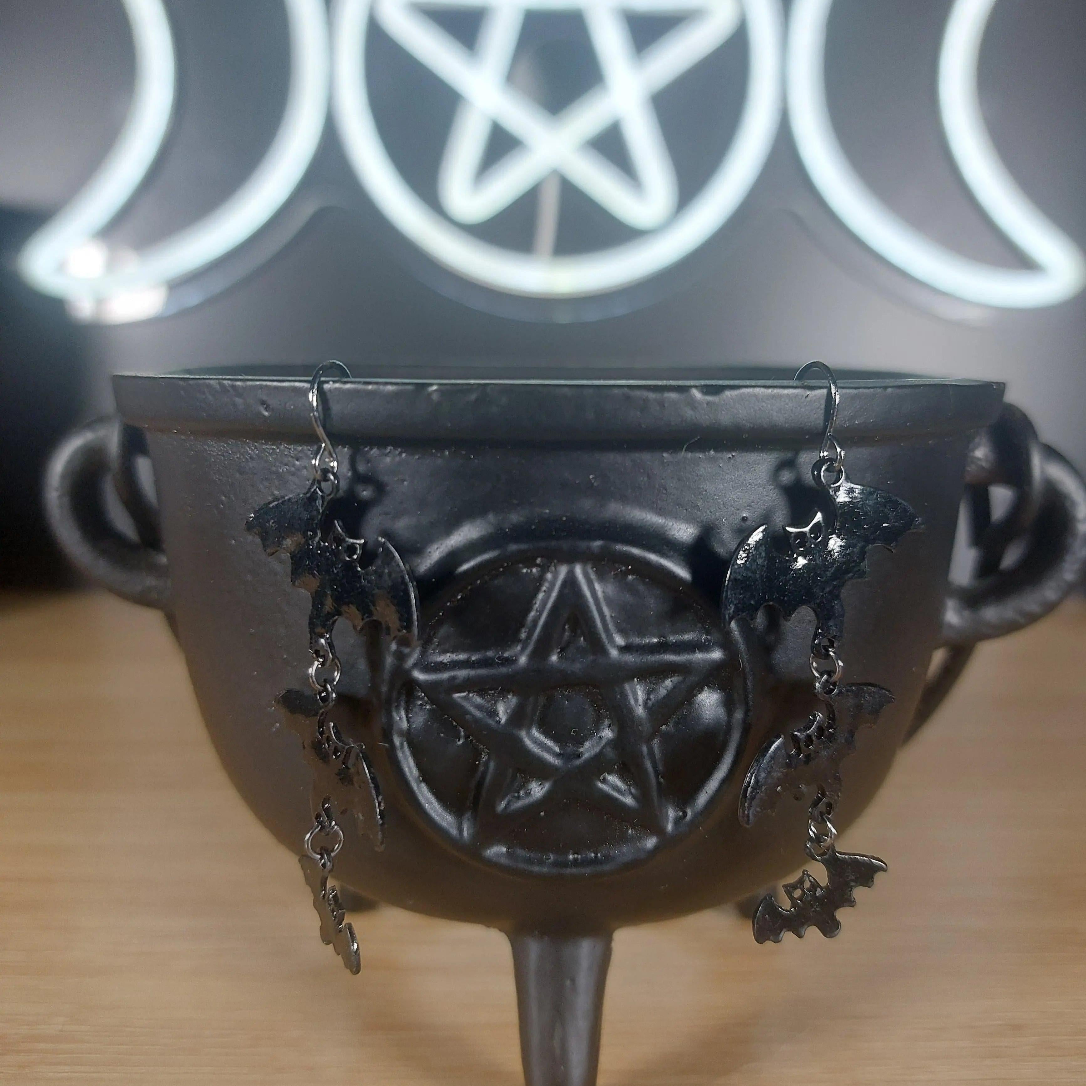 Black metal 3-tier bat dangle earrings hanging from a black cauldron with a pentagram.