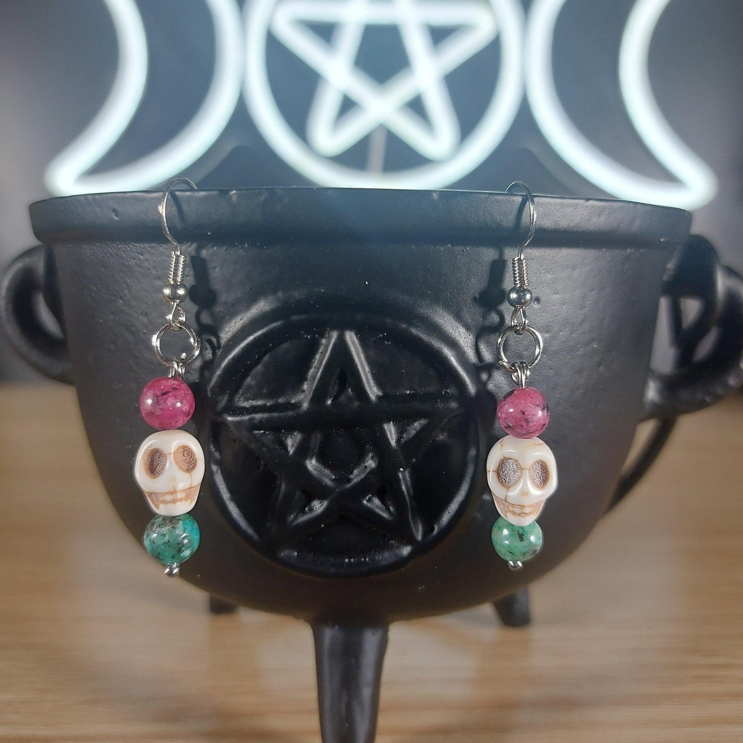 White howlite skull dangle earrings with pink and green glass beads on a black cauldron.