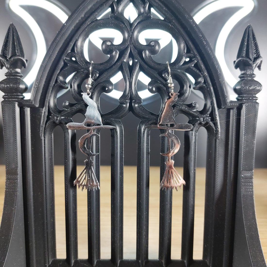 Silver tone broom, witch hat, and crescent moon dangle earrings on a black gothic arch display.