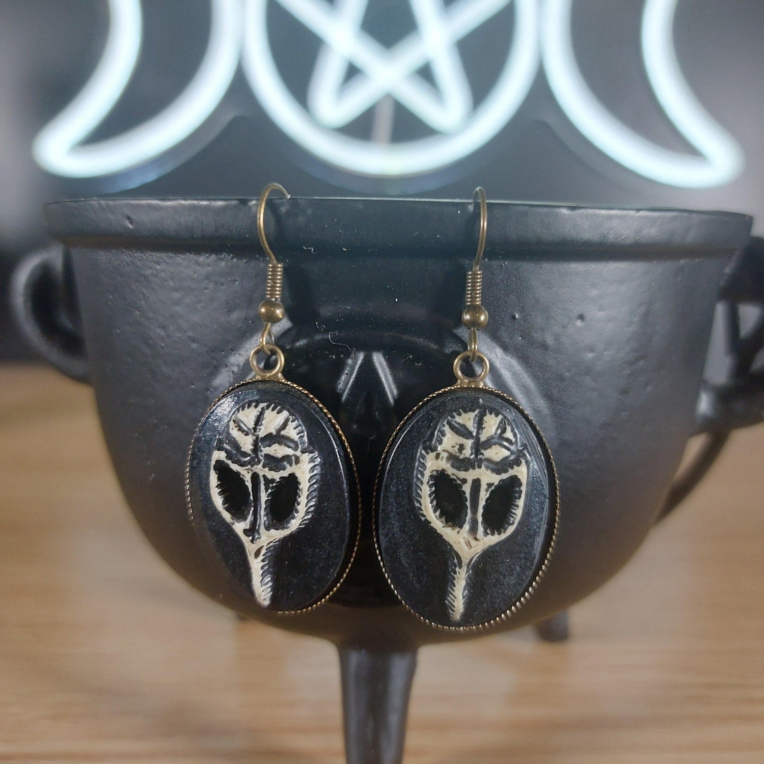 Antique bronze raven skull dangle earrings with black oval settings, hanging from a black cauldron.