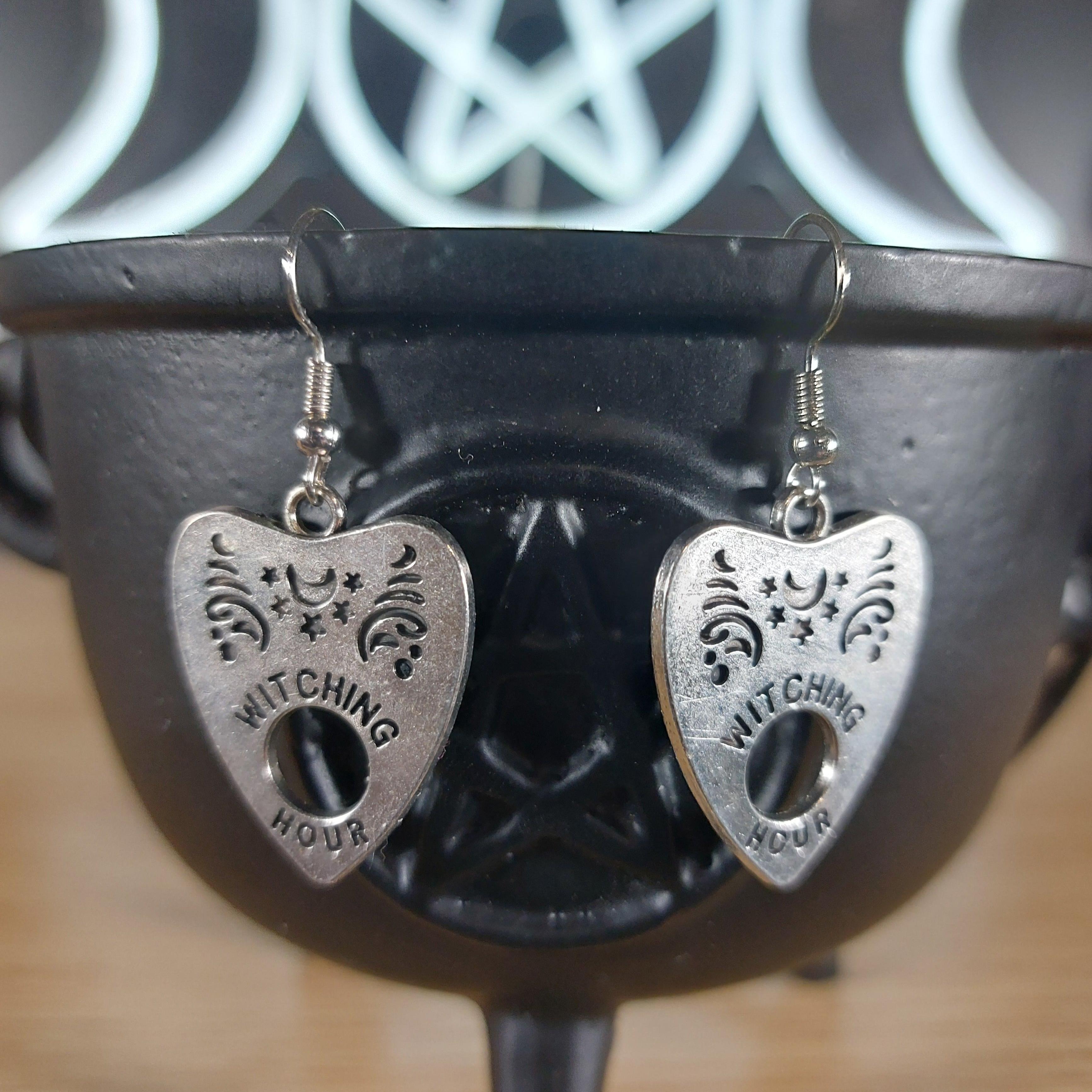 Silver tone Witching Hour Ouija planchette dangle earrings on a black cauldron.
