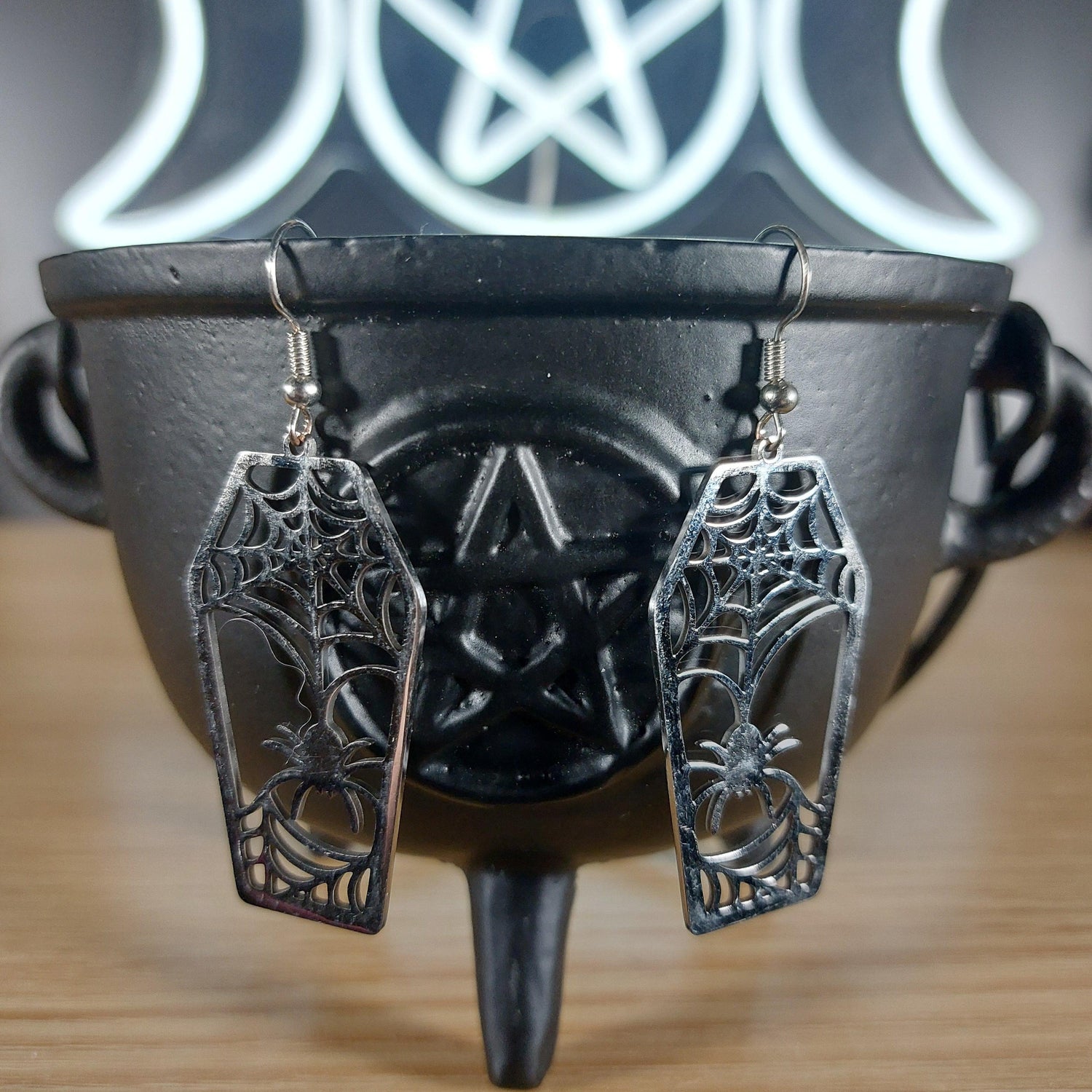 Silver-tone coffin dangle earrings with intricate spider web and spider cutouts, displayed on a black cauldron.