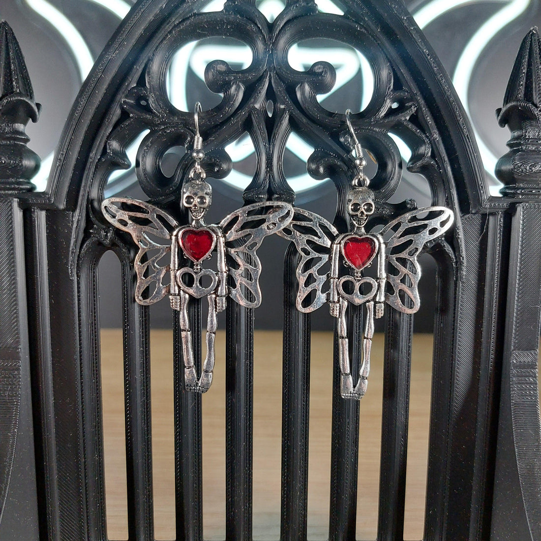 Silver tone skeleton dangle earrings with wings and a red heart, displayed on a black gothic arch.