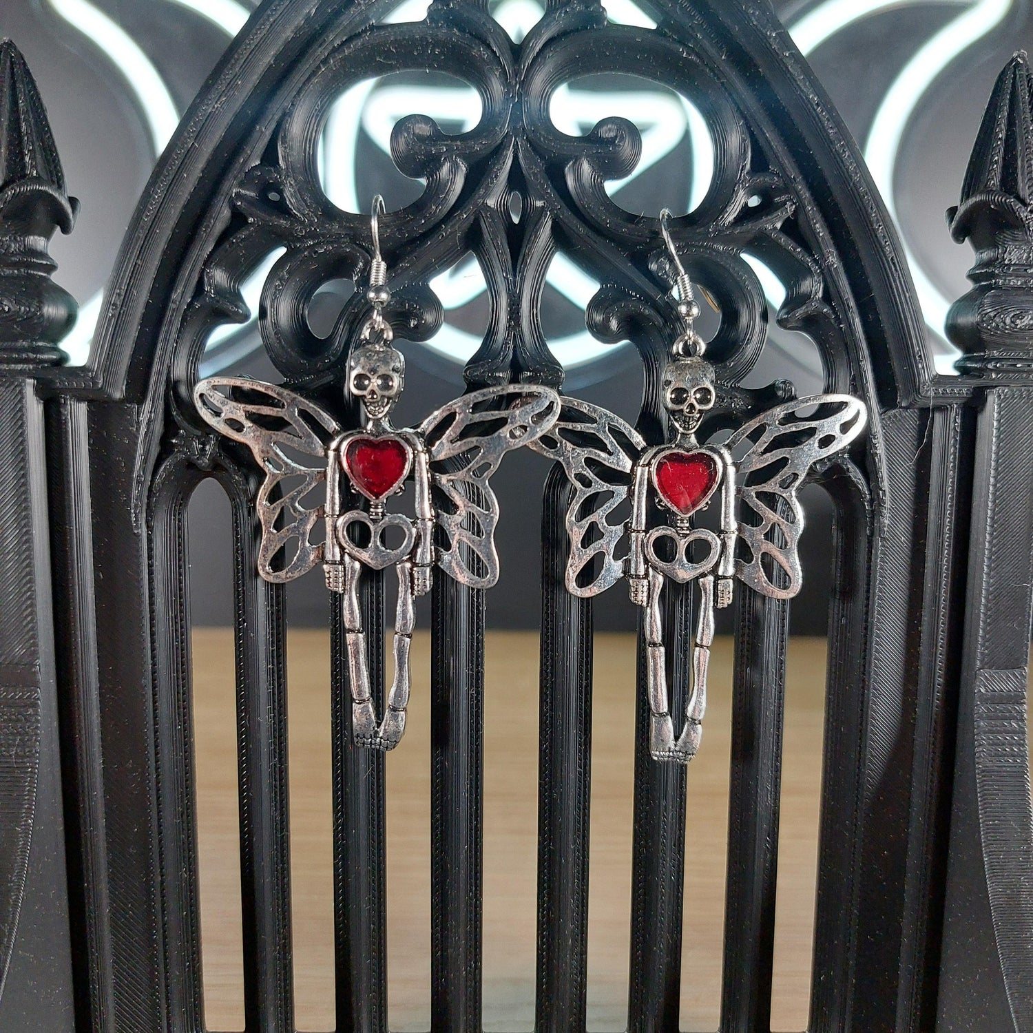 Silver tone skeleton dangle earrings with wings and a red heart, displayed on a black gothic arch.