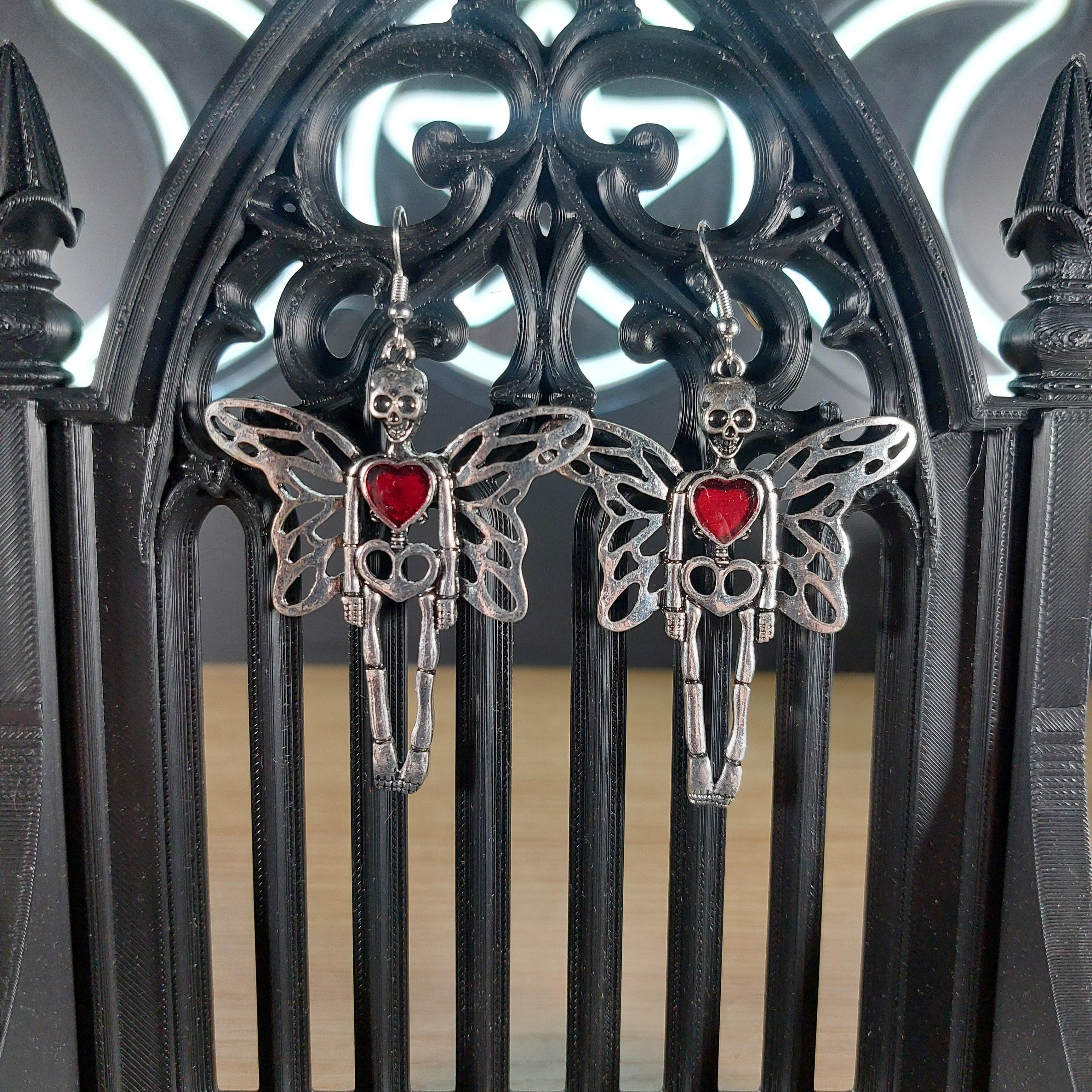 Silver tone skeleton dangle earrings with wings and a red heart, displayed on a black gothic arch.