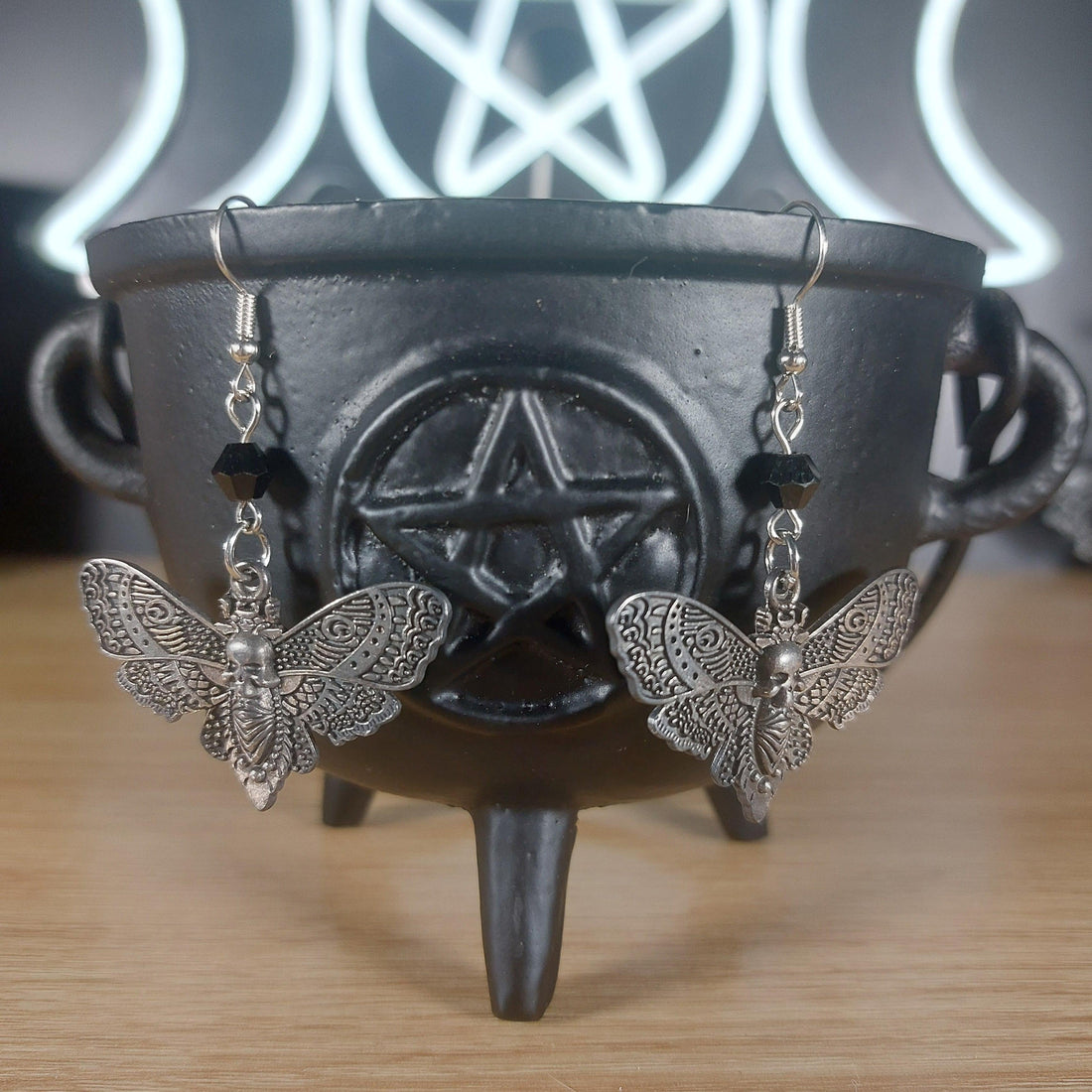 Antique silver death moth dangle earrings with black beads, hanging from a black cauldron.