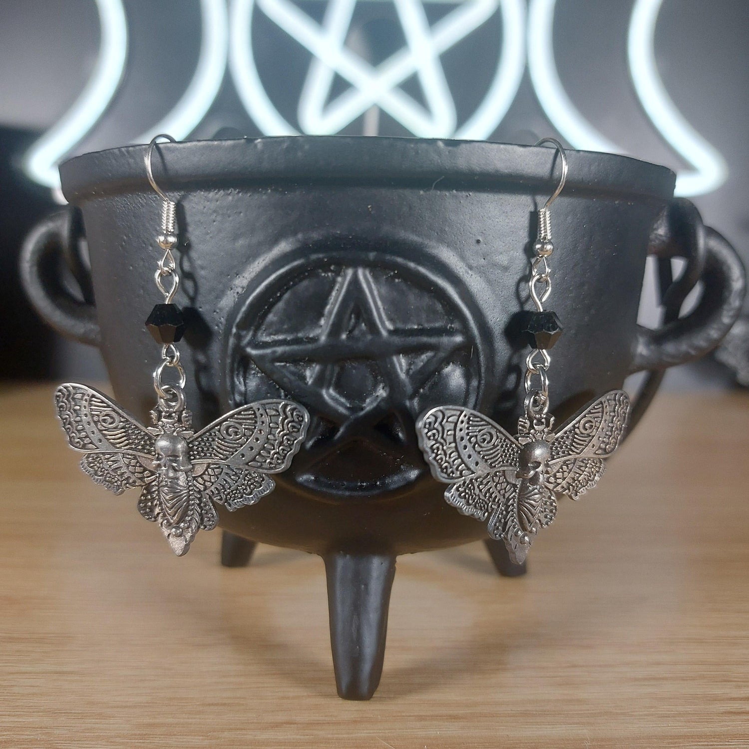 Antique silver death moth dangle earrings with black beads, hanging from a black cauldron.