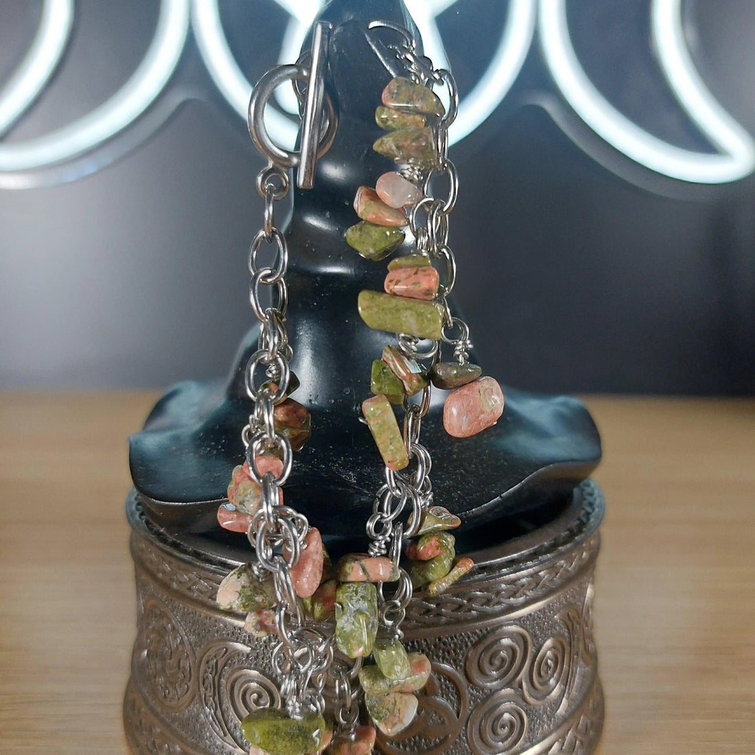 Unakite chip bracelet with green and pink stones on a silver stainless steel chain, displayed on a black stand.