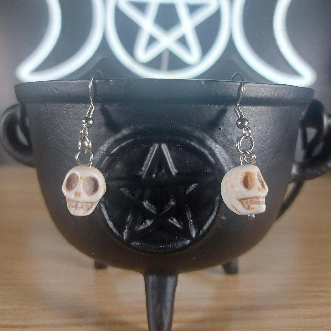 White howlite crystal dangle earrings with skull charms, hanging from a black cauldron.
