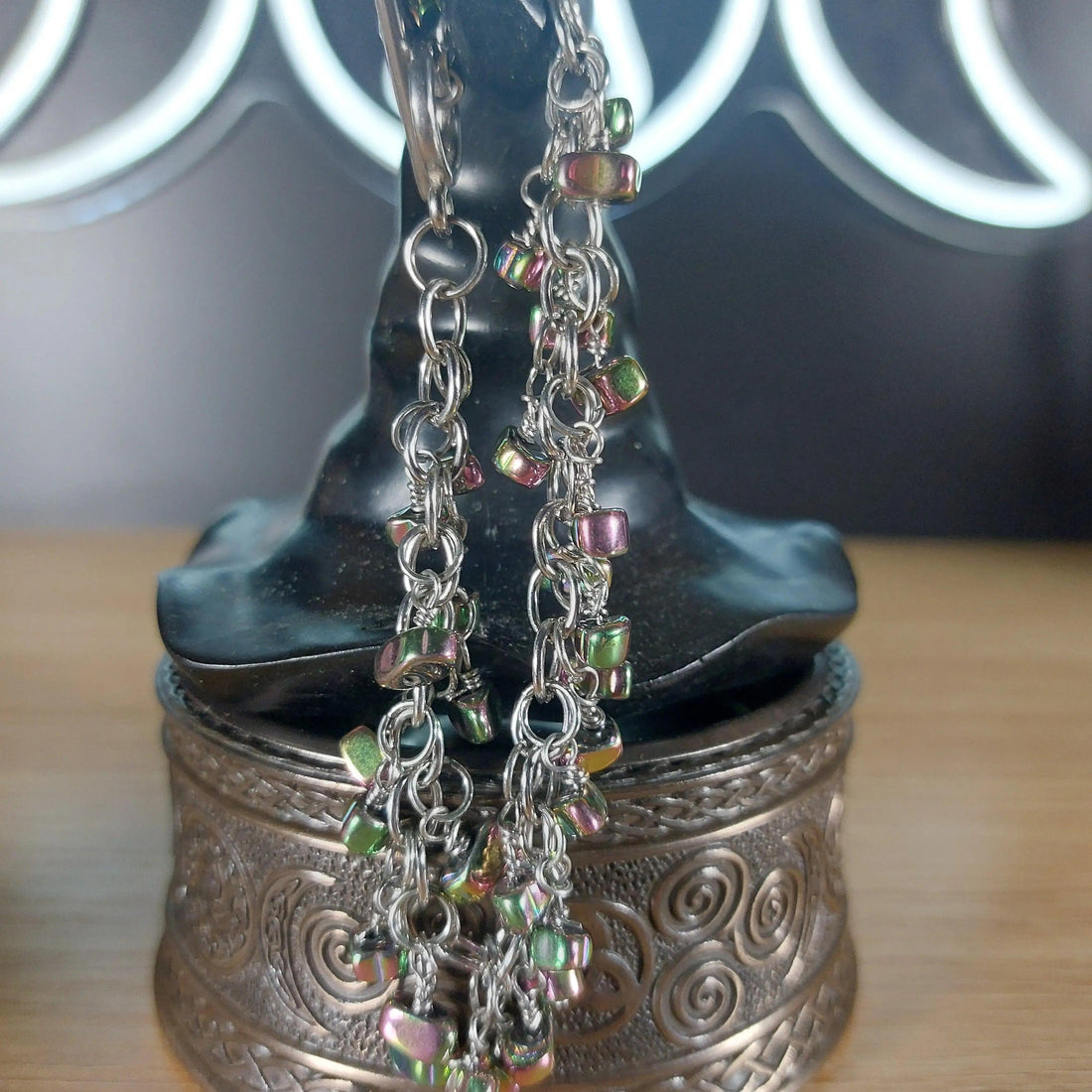 Silver chain dangle earrings with iridescent, multicolored hematite beads on a dark witch hat display.