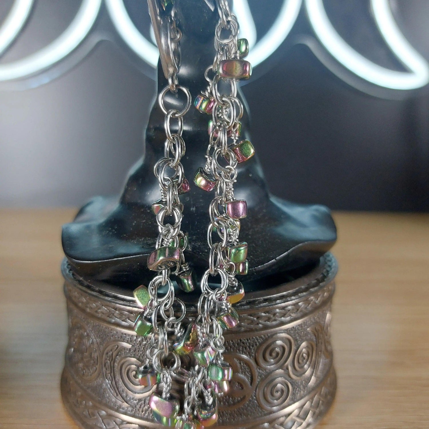 Silver chain dangle earrings with iridescent, multicolored hematite beads on a dark witch hat display.