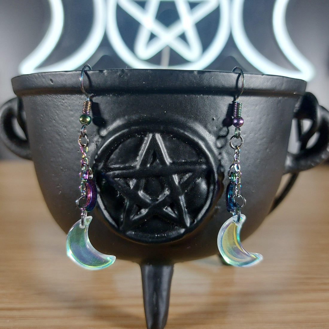 Iridescent glass moon dangle earrings with rainbow beads, hanging from a black cauldron.